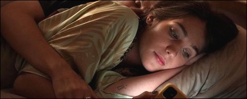 Allegra (Lola Kirke) regarde son téléphone portable au lit et est enlacée par Beth (Soko)